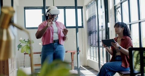 Female coworkers testing virtual reality headset and collaborating in modern glass office