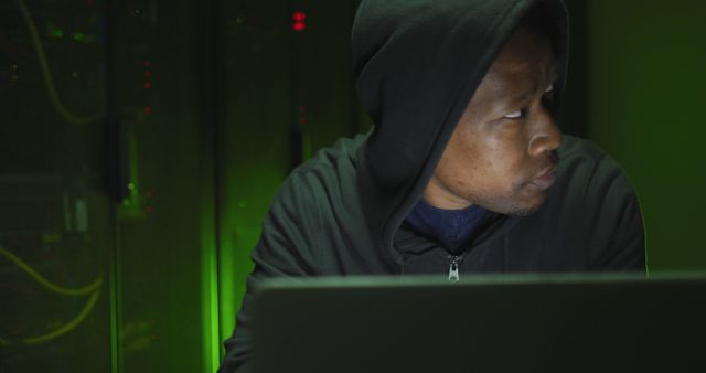 This photo is ideal for articles on cybersecurity, hacking, and digital crimes. It can be used in blogs, news reports, or promotional materials for cybersecurity services. The image depicts a male hacker focused on his work in a dark, green-lit environment, adding to the mysterious and secretive atmosphere.