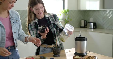 Diverse friends preparing green herb paste in modern kitchen pouring blender into mortar
