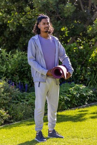 Man holding yoga mat in sunny garden wearing gray tracksuit and trainers preparing for yoga