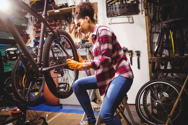 Woman repairing bicycle in a cluttered garage workshop. Useful for topics related to DIY, bicycle maintenance, mechanical repair, and home workshops. Ideal for articles, tutorials, instructional guides, and advertisements for cycling gear or repair services.