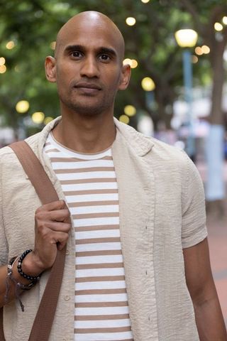 African man standing with shoulder strap wearing striped tee and bracelets on tree-lined street