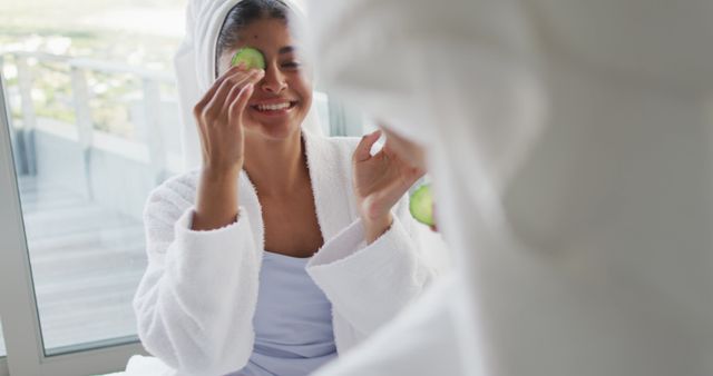 Image of happy diverse female friends in robes having fun with cucumber slices. Friendship, taking care of yourself and beauty concept.