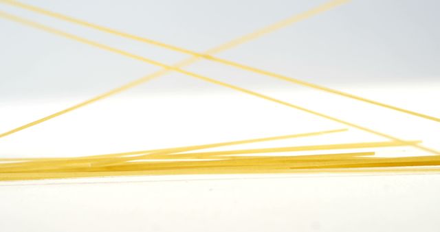 Uncooked spaghetti noodles are scattered and overlapping against a white background, with copy space. Their yellow hue and thin, elongated shapes create an abstract, minimalist aesthetic.