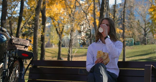 Man sitting on a park bench surrounded by autumn foliage, enjoying a warm coffee and sandwich. Scene suggests leisure and outdoor break during a cool autumn day. Useful for themes around daily routine, casual living, outdoor activities, and relaxation concepts.