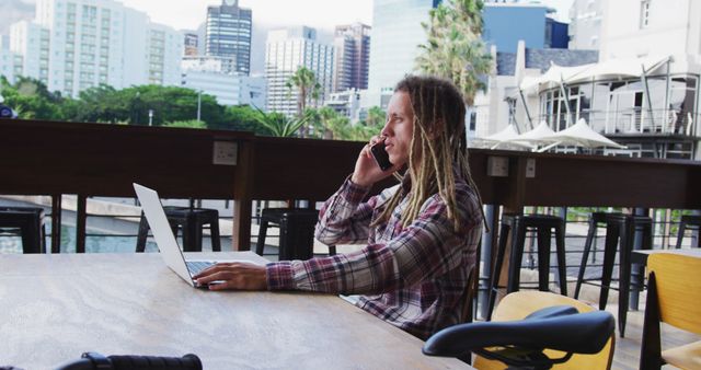 Biracial man with dreadlocks sitting at table outside cafe using laptop and smartphone. digital nomad, out and about in the city.