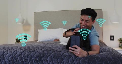 Smiling man petting black dog on bed using smartphone with WiFi icons for smart living