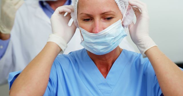 Female healthcare worker wearing blue scrubs and gloves is seen adjusting her face mask for hygiene and protection. This image can be used for health and safety campaigns, hospital or clinic promotional materials, medical presentations, and COVID-19 related information graphics.