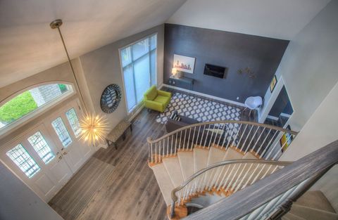 This image showcases a modern living room featuring a stylish spiral staircase, bringing an elegant architectural element to the space. The room benefits from ample natural light streaming in through large windows. Minimalist decor includes a green sofa and a distinctive geometric rug that complements the grey walls. The ambient lighting from the modern chandelier adds to the room's sophisticated atmosphere. Ideal for interior design inspirations, real estate visuals, and home decor themes.