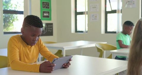 Child using tablet at desk while classmates learning with technology in bright classroom