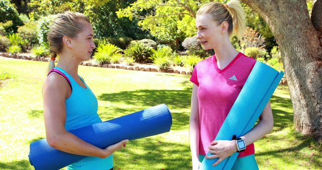 Two women are standing on grass in a park, holding yoga mats and smiling at each other. They appear to be discussing workout plans or catching up after an exercise session. The scene suggests friendship and a healthy lifestyle, making it ideal for content related to fitness, wellness, socializing, or outdoor activities.