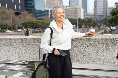 Smiling mature woman leaning on city railing holding coffee and phone, urban commuter