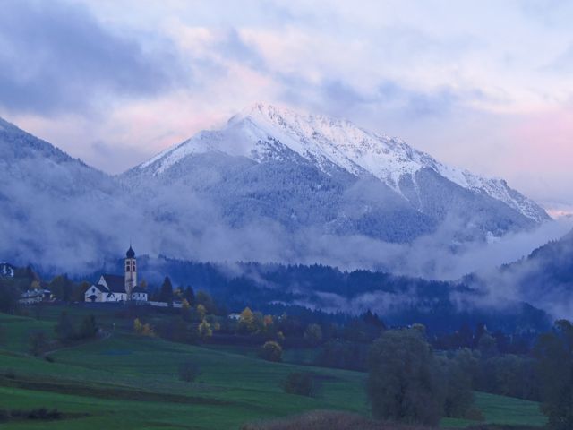 This serene scene features a small countryside church at the base of a snow-covered mountain engulfed in early morning mist. The tranquility of the setting conveys peace and isolation, making it perfect for use in designs emphasizing nature's beauty, calm moments, or depicting idyllic rural life. Ideal for posters, landscapes depicting crisp mornings, travel promotions, and inspirational contexts.