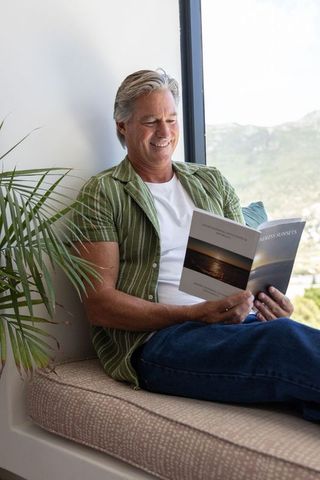 Mature man relaxing on sunlit window seat reading paperback with coastal view and plant
