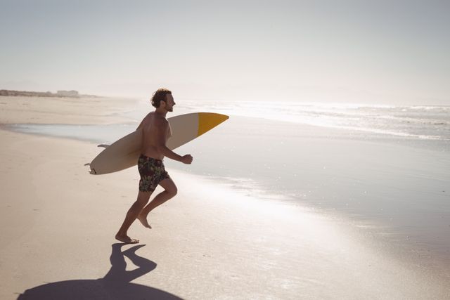 Shirtless man running along the beach while holding a surfboard on a sunny day. Ideal for use in travel brochures, fitness and lifestyle blogs, surfing magazines, and advertisements promoting beach vacations and outdoor activities.