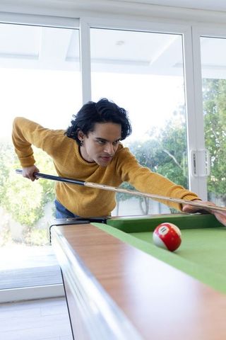 Young man leaning over pool table aiming shot with cue stick in bright home interior