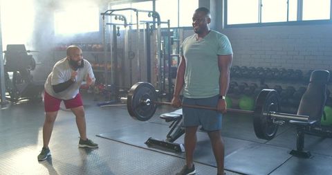 Man deadlifting heavy barbell with coach spotting and encouraging in sunlit commercial gym