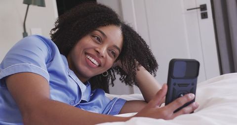 Smiling african woman reclining in light blue satin pajamas using smartphone on cozy bed