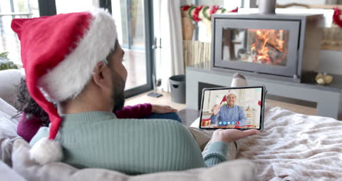 A happy couple wearing Santa hats engages in a virtual video call with a family member, seen on a tablet screen. The seasonal atmosphere is enhanced by a cozy fireplace in the background, contributing to a warm and festive environment. This scene is ideal for illustrating concepts of technology connecting families during the holidays. It can be used for promotions related to video chat services, holiday marketing campaigns, or blog posts about celebrating Christmas remotely.