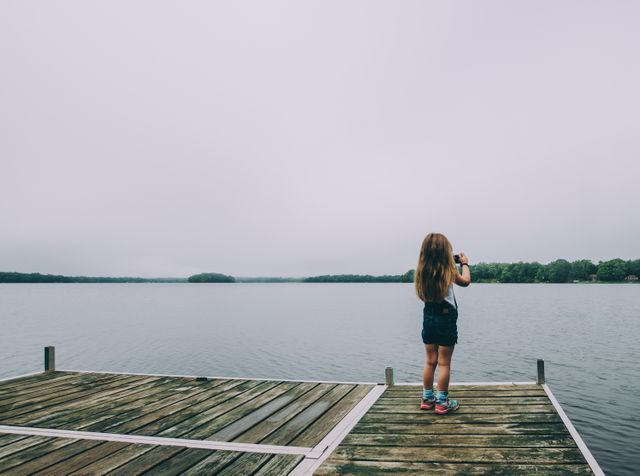 A young girl is standing on a wooden pier taking a photograph of a calm lake with greenery in the background. This image can be utilized for themes of childhood, adventure, summer vacations, nature, tranquility, and photography as a hobby. Ideal for use in lifestyle blogs, travel magazines, educational content, and promotional materials for cameras and outdoor gear.