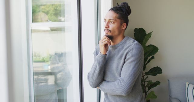Thoughtful biracial man with dreadlocks in hair bun looking out of window in living room. domestic lifestyle, spending time alone at home.