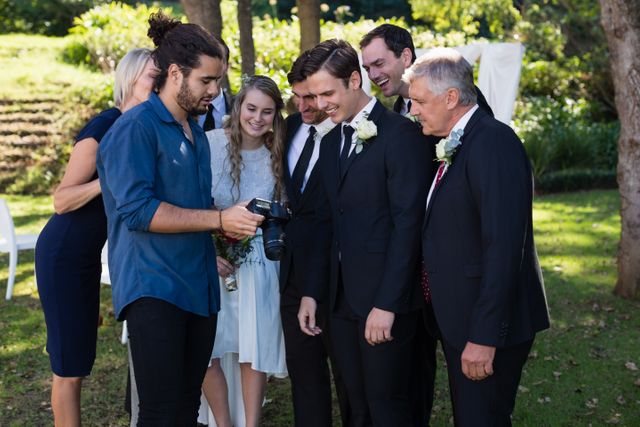Photographer showing wedding photos to bride, groom, and guests in a park. Ideal for use in wedding planning materials, photography portfolios, and articles about wedding photography. Highlights joy and celebration in a natural outdoor setting.