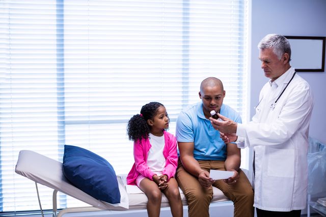 Doctor prescribing medicine to father and daughter in clinic. Father and daughter sitting on examination table while doctor explains medication. Useful for healthcare, medical advice, family health, pediatric care, and patient consultation themes.