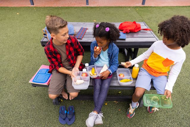 Two diverse boys and a girl eating packed lunches on a bench, talking in elementary schoolyard. Education, health, childhood and learning concept.