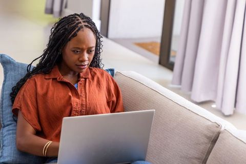 Adult woman with braids working on laptop on sofa in bright minimalist living room
