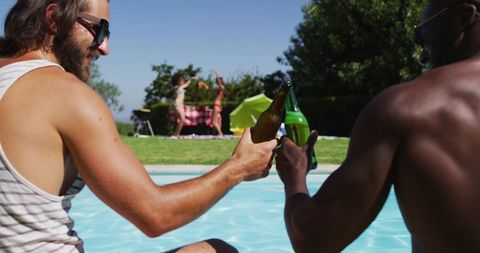 Friends clinking beer bottles by backyard pool celebrating sunny summer picnic and bbq
