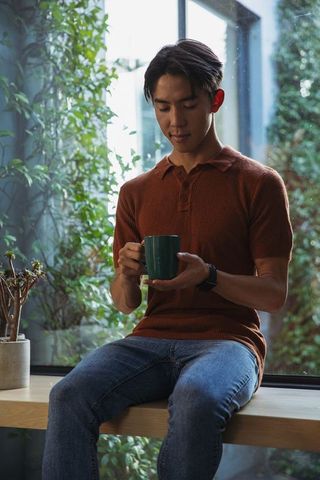 Asian man sitting on window bench cradling green mug, enjoying quiet moment with plants