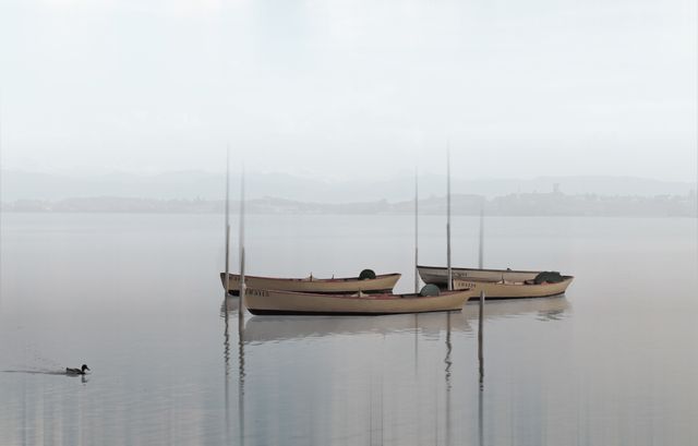 Boats floating on a calm lake during a misty morning. Serene and tranquil atmosphere with fog gently enveloping the water and soft reflections of the boats. Perfect for use in nature-themed projects, promoting calmness, relaxation, vacation advertisements, or background images.