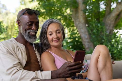 Mature interracial couple relaxing on outdoor sofa scrolling smartphone in sunlit garden