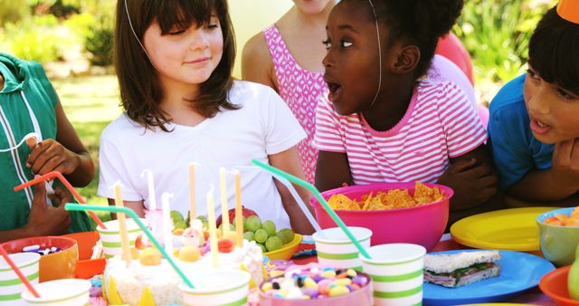 A diverse group of children is gathered around a table at an outdoor birthday party, with copy space. Excitement and joy are evident on their faces as they celebrate the special occasion with snacks and birthday cake.