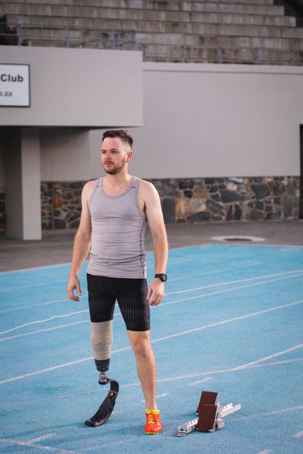 Caucasian disabled male athlete with prosthetic leg training, standing on runway. professional runner training at sports stadium.