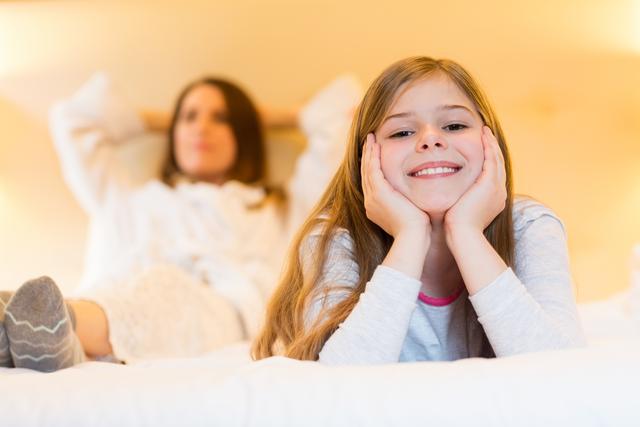 Young girl smiling while lying on bed with mother in background. Perfect for family lifestyle, home comfort, and childhood happiness themes. Ideal for use in parenting blogs, family-oriented advertisements, and home decor promotions.