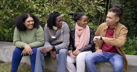 Diverse friends laughing on park bench outdoors, chatting and checking watch, wearing scarves