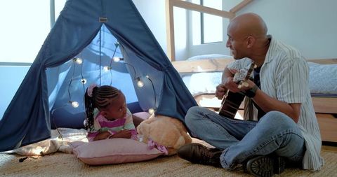 African american father playing guitar while daughter lying in cozy blue tent in bedroom