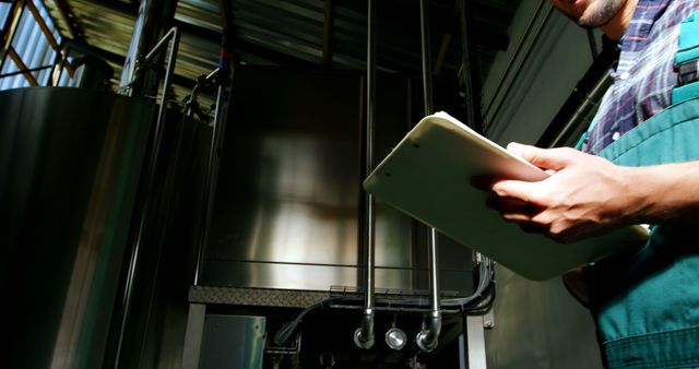A technician in an industrial plant is holding a clipboard while inspecting machinery. The person is focusing on quality control and maintaining equipment at a manufacturing facility. Useful for illustrating concepts of factory work, engineering processes, industrial inspections, and maintenance tasks in promotional materials, websites, and educational resources.