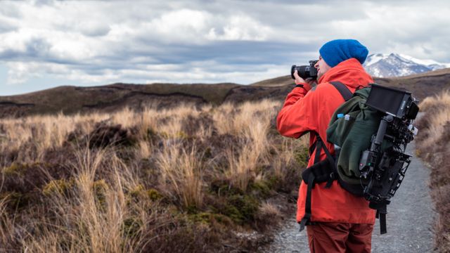 Photographer wearing red jacket standing on trail, taking photo of scenic outdoors in wilderness. Ideal for content on nature photography, adventure travel, and outdoor activities. Perfect for campaigns promoting tourism, photography equipment, and adventurous lifestyles.