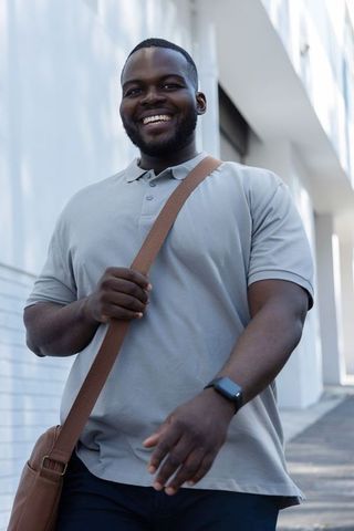 Smiling african american man walking with crossbody bag and smartwatch in urban daylight