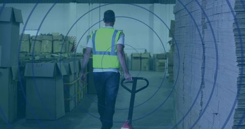 Warehouse worker pulling pallet jack through aisle with stacked cardboard and neon vest