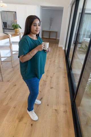 African american woman holding mug gazing through sliding glass doors in modern open-plan home