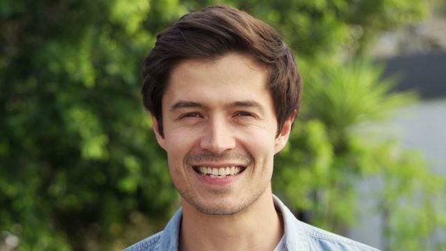 A young Caucasian man smiling while socializing outdoors, with lush greenery in the background. His casual denim shirt enhances the relaxed, friendly vibe. This can be used for lifestyle commercials, blog posts on social interactions, or advertisements for casual and outdoor clothing brands.