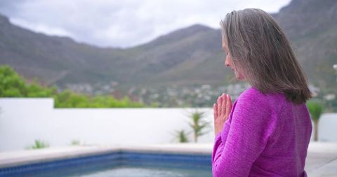 Mature woman meditating poolside on mountain terrace in magenta sweater