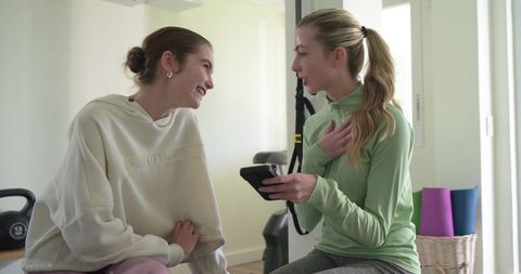 Women laughing and sharing smartphone while resting in bright home gym with kettlebell