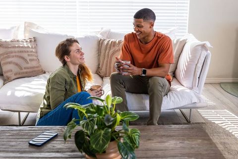 Sunlit mixed-race couple relaxing and chatting over coffee mugs in cozy modern living room