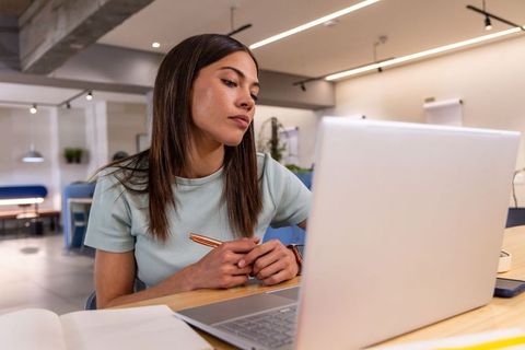 Young woman working on laptop in modern coworking lounge holding pen and taking notes