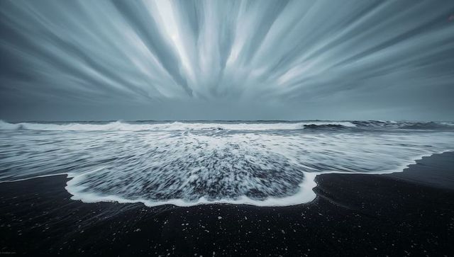 Surging foamy wave on black sand beach with dramatic streaked clouds, long exposure