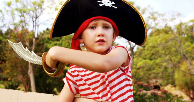 Young boy wearing a classic pirate hat and red striped shirt, holding a toy sword, playing outside. Perfect for promoting children's parties, imaginative play, kids' activities, and pirate-themed events. Suitable for use in advertisements, educational materials, storytelling visuals, and parenting blogs.